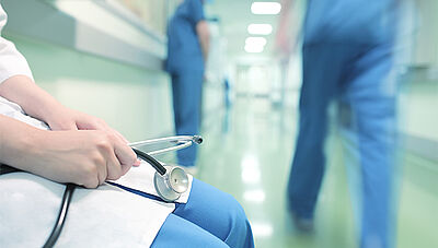Médecin en blouse blanche avec un stéthoscope assis dans un couloir d'hôpital avec du personnel soignant en tenues bleues autour. Conférence Prise en charge des maladies rares : enjeux pour le patient et le système de santé, Executive MBA Santé Dauphine-PSL
